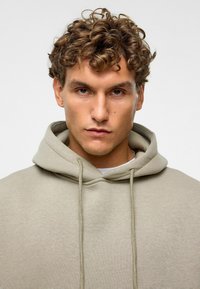 Beige Kapuzenpullover mit Kordelzug, der eine weiche Textur und einen großen Ausschnitt hat. Der Stoff wirkt leicht und lässig.