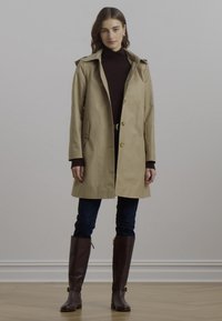 Trench coat beige con colletto, due tasche frontali e chiusura a bottoni. Indossato sopra un dolcevita scuro e abbinato a jeans aderenti e stivali marroni.