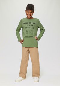 Grünes Langarmhemd mit Text und Grafik eines Basketballfeldes. Kombiniert mit hellkhakifarbenen weiten Hosen und weißen Sneakers. Einfaches Design.