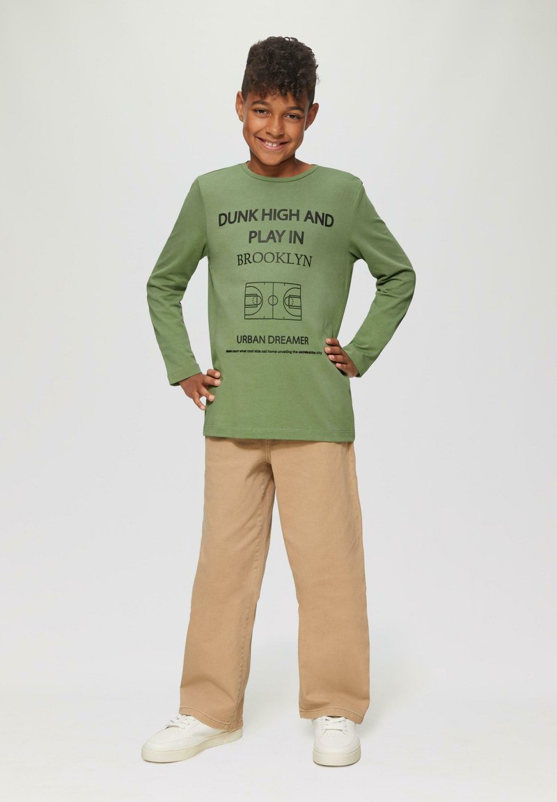 Grünes Langarmhemd mit Text und Grafik eines Basketballfeldes. Kombiniert mit hellkhakifarbenen weiten Hosen und weißen Sneakers. Einfaches Design.