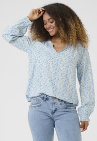 Blumenbluse in Hellblau mit gelben und blauen Blumen; hat einen V-Ausschnitt und lange, geraffte Ärmel. Kombiniert mit hellblauen Jeans.