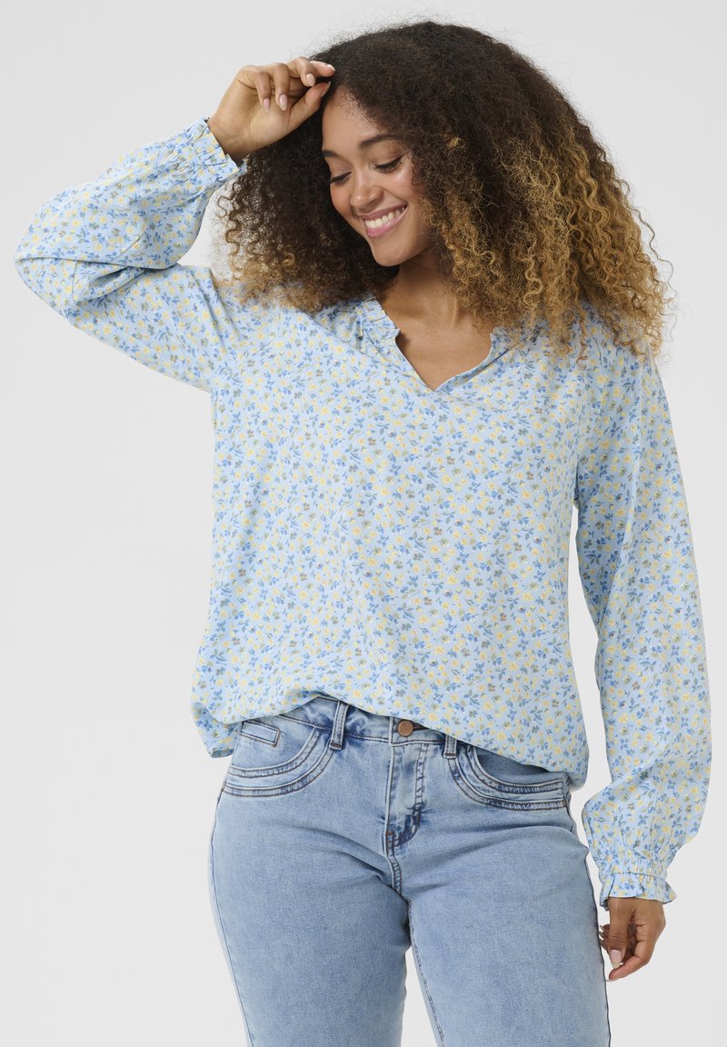 Blumenbluse in Hellblau mit gelben und blauen Blumen; hat einen V-Ausschnitt und lange, geraffte Ärmel. Kombiniert mit hellblauen Jeans.