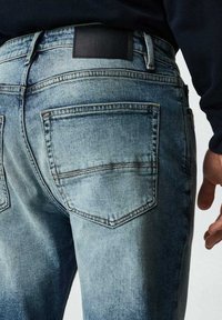Světle modré džíny z denimu s vybledlým vzhledem, vybavené dvěma zadními kapsami a černou koženou nášivkou na pásku. Povrch je hladký s mírným opotřebením.