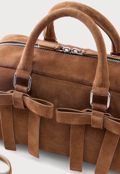 Καφέ τσάντα από σουέτ με στρογγυλεμένες λαβές, ασημένιο φερμουάρ και διακοσμητικά φιόγκους στο μπροστινό μέρος, τοποθετημένη πάνω σε λευκό φόντο.