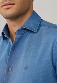 Lichtblauwe denimshirt met een gespreide kraag, knoopsluiting en gestikte accenten. Heeft een subtiele geborduurde logo op de borst.