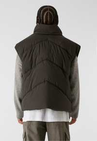 Dunkelgrüne Steppweste mit hohem Kragen, übergroßen Armausschnitten und Zickzack-Steppung. Getragen über einem hellgrauen Pullover und einer weißen Bluse mit mehreren Lagen.