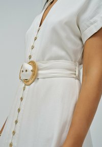 Vestido blanco de camisa con mangas cortas, que presenta un escote en V profundo, detalles de botones y un cinturón ancho acentuado por una gran hebilla redonda en color marrón claro.