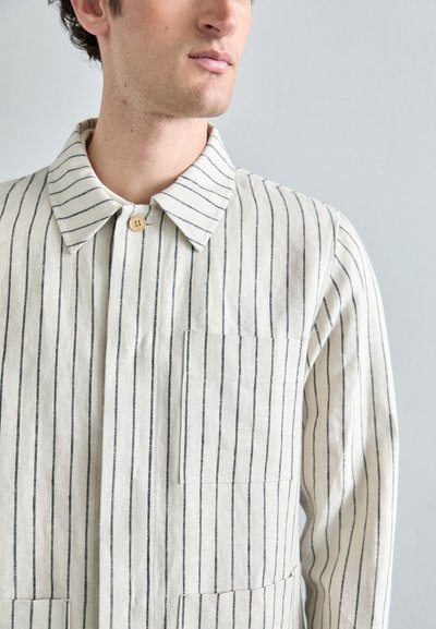 Chemise en lin à rayures, avec un col boutonné, des rayures verticales bleu marine sur un fond beige, et une poche poitrine pour plus de fonctionnalité.