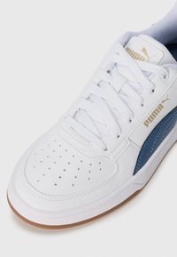 Chaussure de sport blanche avec des accents bleus, tige perforée pour la respirabilité, col rembourré et lacets plats. Présente une semelle en caoutchouc gomme pour l'adhérence.