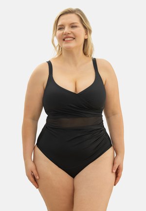 Lächelnde Frau, die einen schwarzen Einteiler mit verstellbaren Trägern und einem Mesh-Detail in der Taille trägt und vor einem schlichten Hintergrund steht.