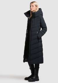 Abrigo puffer largo en azul marino oscuro con cuello alto y cierre de cremallera. Patrón texturizado y acolchado con bolsillos laterales y una silueta moderna.