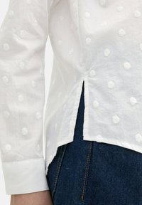 Weiße Baumwollbluse mit strukturiertem Punktmuster, seitlichen Schlitzen und angeschnittenen Ärmeln, kombiniert mit dunklen Jeans.