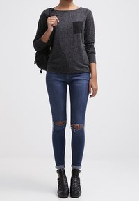 Dunkelgraues Oberteil mit langen Ärmeln und Struktur, ausgestattet mit einer schwarzen Tasche, kombiniert mit zerissenen, dunkelblauen Skinny-Jeans und schwarzen Schnür-Stiefeletten.