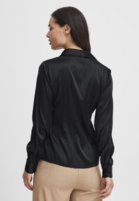 Schwarze Satinbluse mit Kragen, langen Ärmeln und Knopfmanschetten. Der Rücken hat eine glatte Textur und eine leicht taillierte Passform.