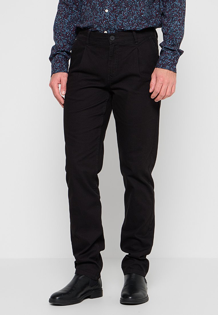 Mann in schwarzen Slim-Fit-Hosen, schwarzen Lederstiefeln und einem dunkel gemusterten Langarmhemd, das vor einem einfarbigen, hellen Hintergrund steht.