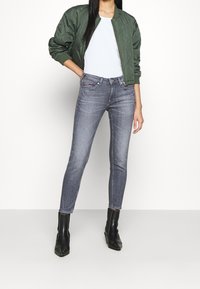 Chaqueta bomber acolchada verde, camiseta ajustada azul clara, jeans ajustados grises y botas negras de tobillo. La chaqueta presenta puños y dobladillo de canalé.