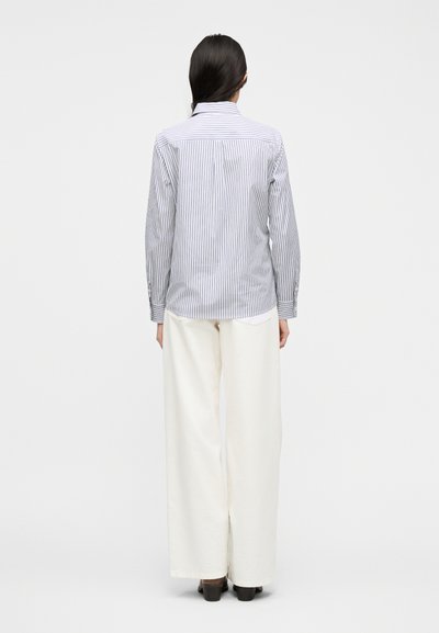 Άτομο με μακριά σκούρα μαλλιά, φορώντας μπλούζα με μακριά μανίκια με μπλε ρίγες, φαρδιά λευκή παντελόνια και σκούρα παπούτσια, στέκεται με την πλάτη στραμμένη σε λευκό φόντο.