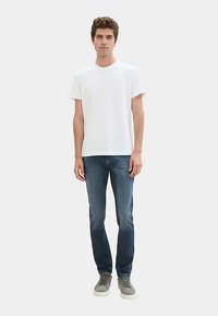 Camiseta blanca de algodón con cuello redondo, combinada con vaqueros de mezclilla azul oscuro. El modelo lleva zapatillas grises. Diseño sencillo y ajuste relajado.