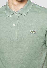 Homme portant un polo vert clair avec deux boutons blancs et un petit logo de crocodile vert sur la poitrine gauche.