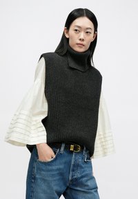 Vrouw draagt een zwart gebreid mouwloos coltrui-vest over een witte blouse met wijde mouwen en blauwe spijkerbroek, handen in de zakken, kijkt zijwaarts.