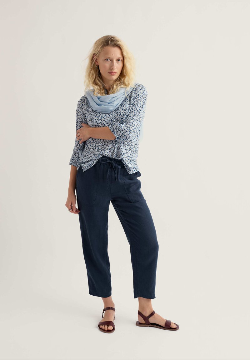 Femme debout, les bras croisés, portant un chemisier à fleurs, une écharpe bleu clair, un pantalon à cordon de serrage marine et des sandales marron.