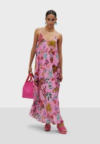 Robe maxi florale rose à bretelles spaghetti, ornée de motifs botaniques multicolores. La modèle tient un sac à main rose et porte des sandales à plateforme.