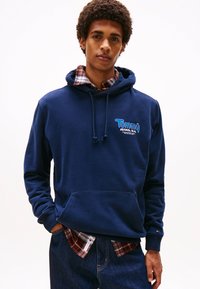 Navy blue hoodie made of cotton, featuring a front pocket and a drawstring hood. Underneath, a red and navy plaid shirt is visible.
