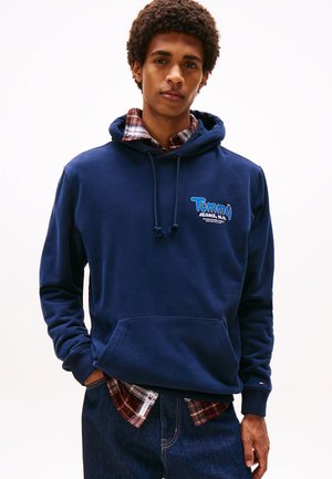 Marineblauwe hoodie van katoen, met een voorvak en een capuchon met trekkoord. Daaronder is een rood en marineblauw geruit shirt zichtbaar.