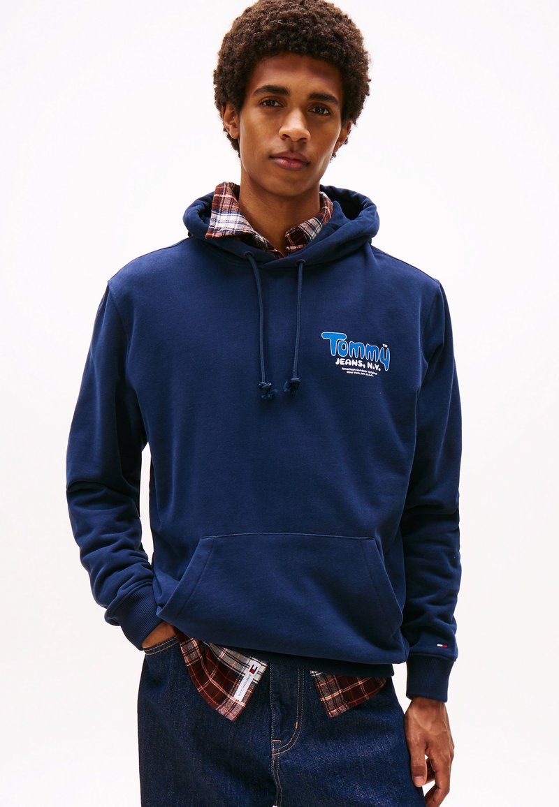 Navy blue hoodie made of cotton, featuring a front pocket and a drawstring hood. Underneath, a red and navy plaid shirt is visible.