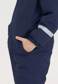 Mørkeblå vandtæt overall med elastiske manchetter, sidelommer og reflekterende sølvdetaljer på ærmerne. Glat tekstur, skræddersyet pasform.