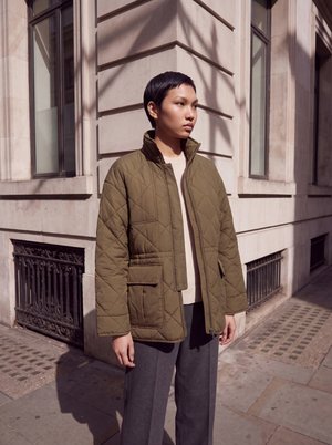 Une personne porte une veste matelassée marron et un pantalon foncé sur un trottoir ensoleillé, à côté d’un bâtiment en pierre.