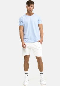 Lyseblå bomulds t-shirt med rund halsudskæring, parret med hvide shorts. Modellen har hvide sneakers og stripede sokker på, stående mod en ensfarvet baggrund.