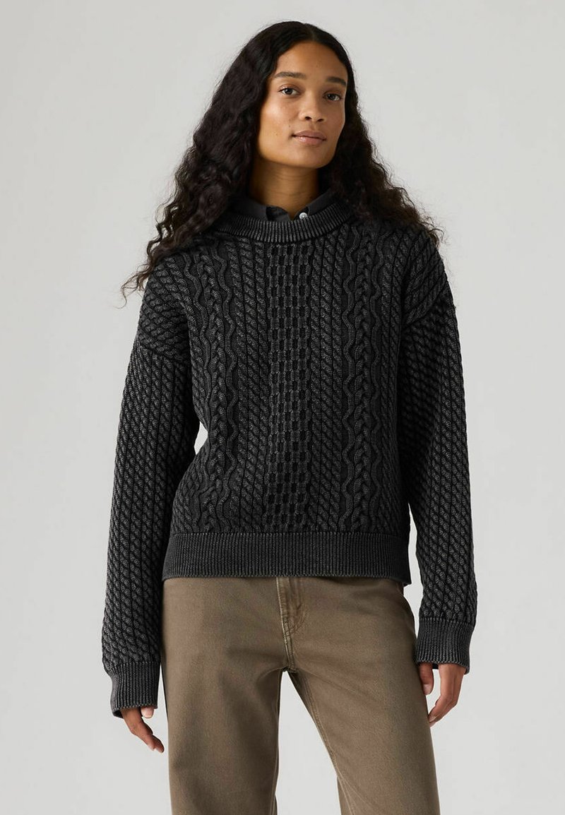 Jersey negro de punto con patrones de trenzas texturizadas, puños anchos acanalados y diseño de cuello alto. La modelo lleva pantalones kaki.