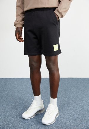 Shorts de algodón negros con una cinturilla elástica, que presentan un destacado parche de logo en color verde lima. Se llevan con zapatillas blancas y calcetines.