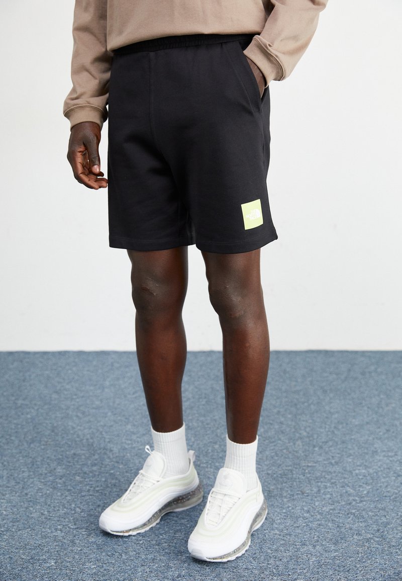 Shorts de algodón negros con una cinturilla elástica, que presentan un destacado parche de logo en color verde lima. Se llevan con zapatillas blancas y calcetines.