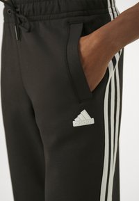 Čierne športové nohavice vyrobené z mäkkého materiálu, s bielymi pruhmi po stranách a vyrazeným logom Adidas na stehne.