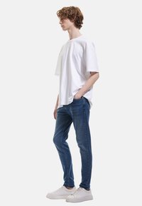 Camiseta blanca de gran tamaño con un corte holgado, combinada con jeans ajustados azules y zapatillas blancas, creando un look casual con un detalle mínimo.