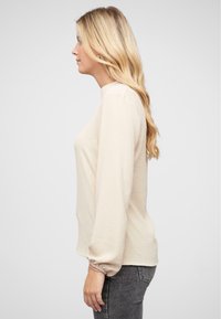 Top a maniche lunghe di colore crema in un tessuto texture, con scollatura rotonda e polsini elastici. Abbinato a jeans aderenti grigio scuro.
