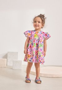 Vestito colorato in tessuto rosa con disegni di faccine sorridenti, stelle e animali. Caratterizzato da maniche svolazzanti e vita arricciata. Indossato con sandali multicolore.