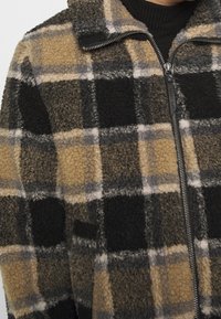 Chaqueta de forro polar con un patrón de cuadros en tono marrón claro, negro y gris. Cuenta con un cuello alto y un cierre con cremallera en el frente. Textura suave visible.