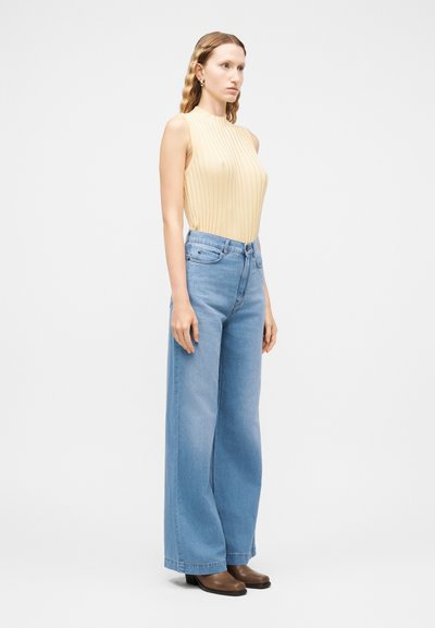 Femme aux cheveux ondulés portant un haut beige côtelé sans manches, un jean bleu taille haute à jambes larges et des chaussures marron à talons, debout de profil.