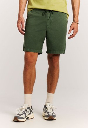 Man draagt olijfgroene korte broek, lichtgeel T-shirt, witte sokken en grijs-witte sneakers, staand tegen een effen achtergrond.