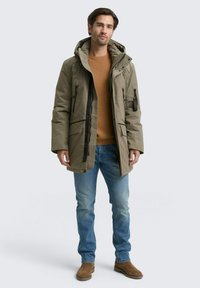 Khaki parka s kapucňou, so zipsom vpredu a viacerými vreckami, nosený cez hnedý sveter a modré džínsy. Hladká textúra látky.