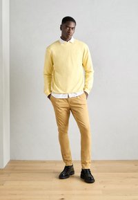 Un pull jaune à manches longues sur une chemise blanche à col, associé à un pantalon slim beige et des chaussures noires, sur un fond de sol en bois clair.
