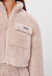 Kremowa kurtka z polaru z wysokim kołnierzem, zapinana na zamek błyskawiczny, z kieszenią na piersi. Miękka w dotyku, o krótkim fasonie.