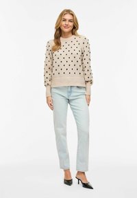Femme portant un pull beige à pois noirs, un jean bleu clair et des mules noires à talons, debout devant un fond blanc.