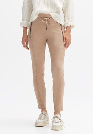 Personne portant un pull crème côtelé, un pantalon beige à cordon avec des fermetures éclair devant et des baskets blanches, debout devant un fond uni.