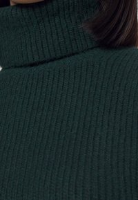 Dunkelgrüner, gerippter Rollkragenpullover mit weicher Textur. Der Stoff weist vertikale Streifen auf, die Wärme und Komfort betonen.