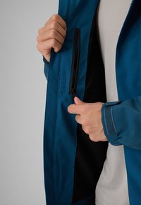 Des mains qui ferment la poche intérieure zippée d'une veste bleue, montrant les détails du tissu et de la fermeture de la veste sur un fond clair.