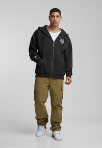 Sort zip-up hættejakke med en frontlomme, prydet med hvidt logo på brystet; parret med olivengrønne cargo-bukser og hvide sneakers.